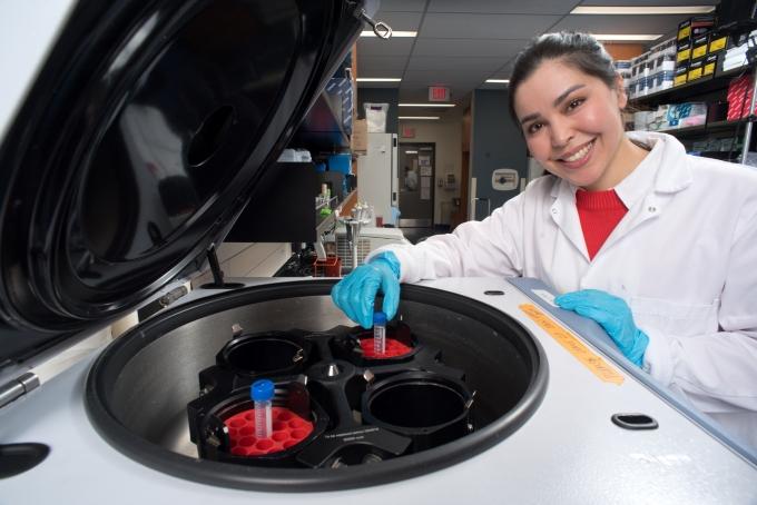 Dr. Fazel wears a lab coat and gloves, smiling as she reaches into a centrifuge to pull out a test tube