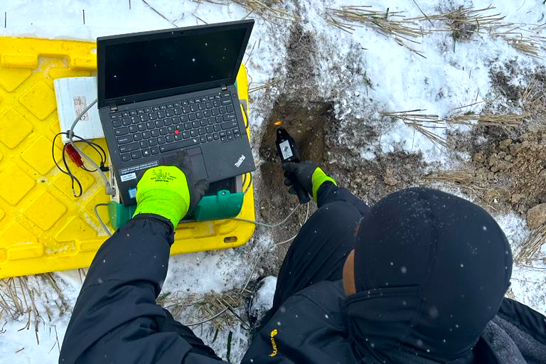 A researcher sits on soil lightly covered in snow, working on a laptop