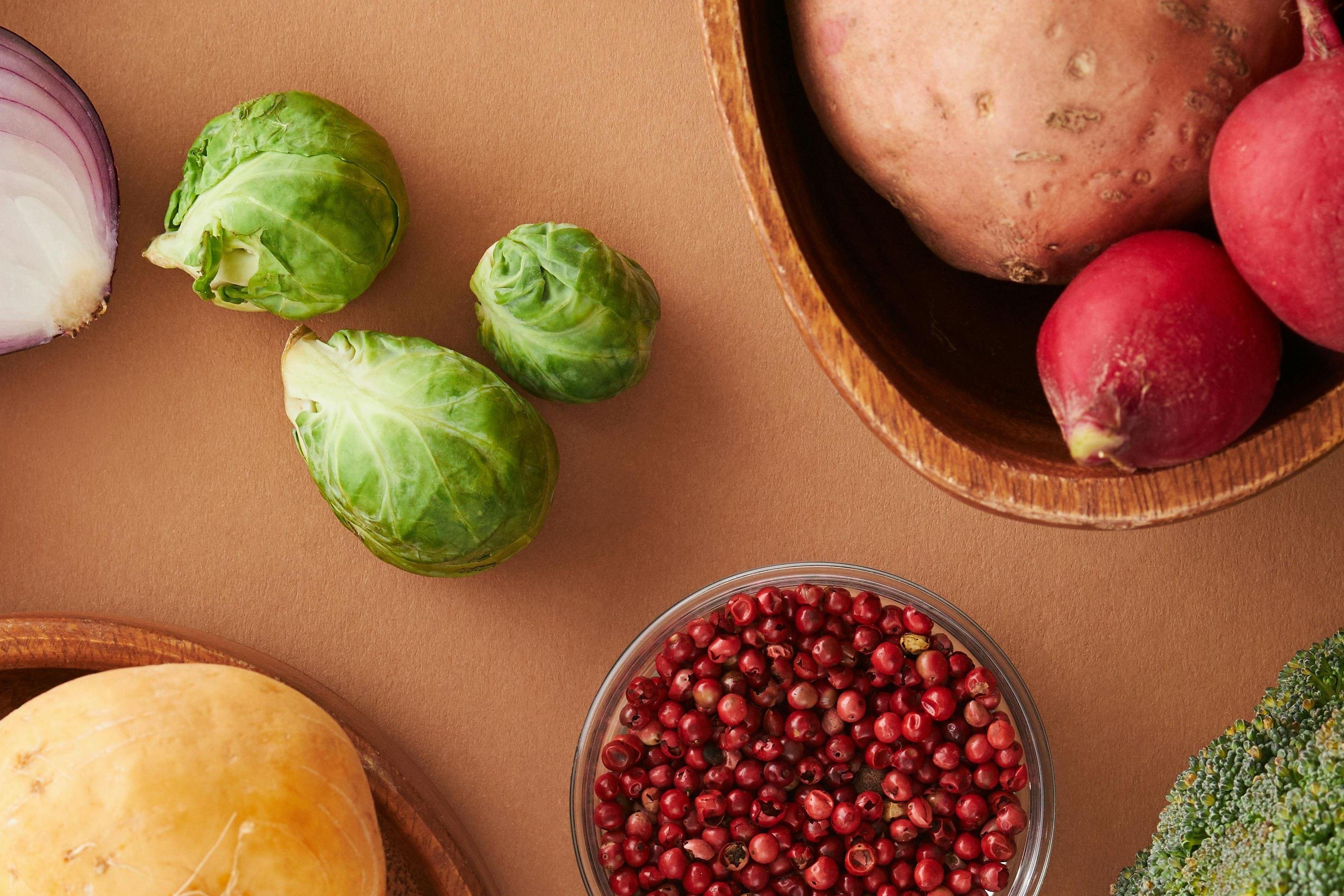Pieces of raw foods, including brussel sprouts, potatoes and pink peppercorn, sitting on a brown table.