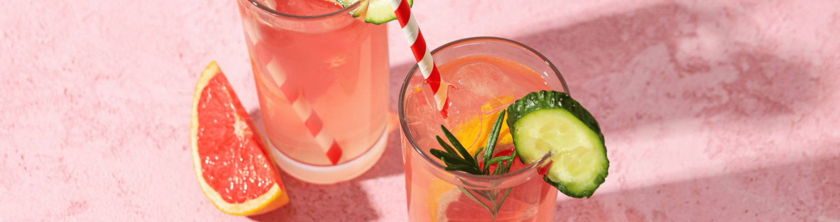 Parts of 2 glasses with pink cocktails in it with grapefruit and cucumber nearby, on a pink table.