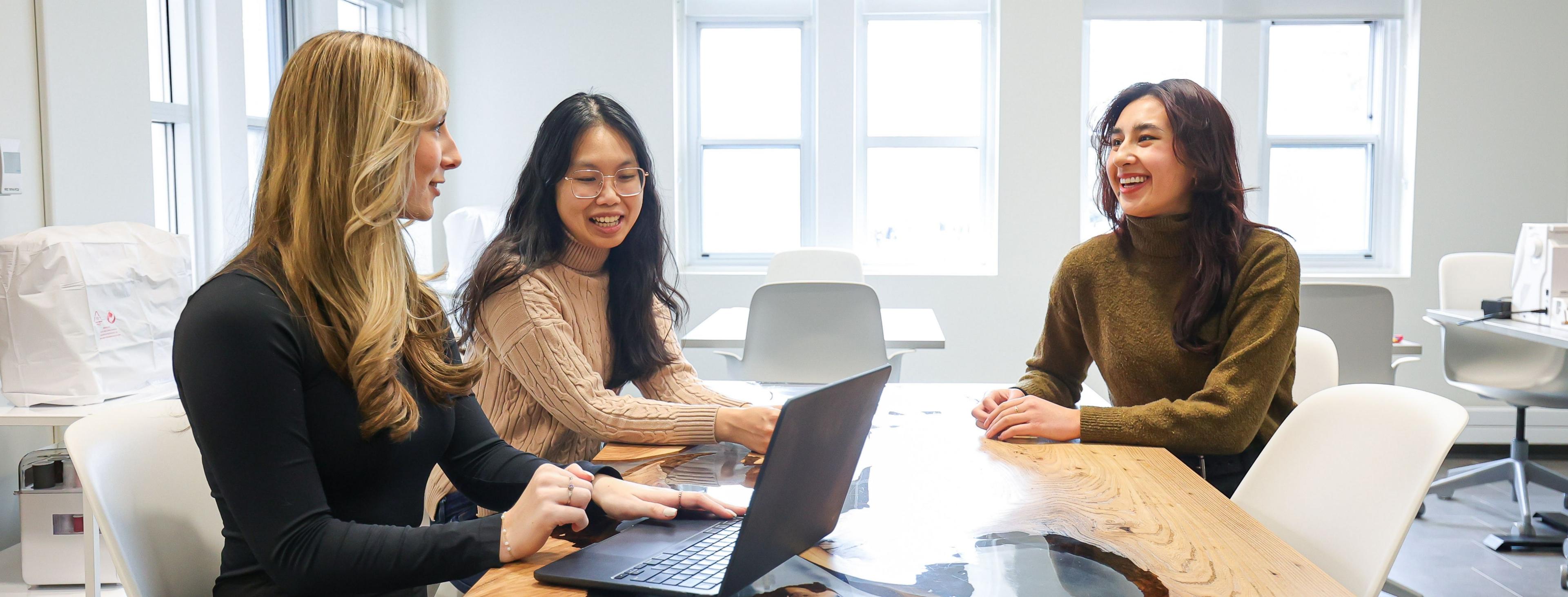 Three students chat at a worktable with a laptop, in a brightly lit office space.